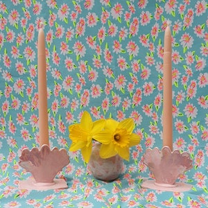 May include: Two pink seashell candle holders with peach candles, and a small vase of yellow daffodils. The background is a floral pattern of white and pink flowers on a turquoise background.