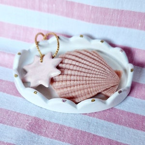 May include: A white ceramic dish with a scalloped edge and gold accents, featuring a pink seashell and a pink star-shaped ornament.