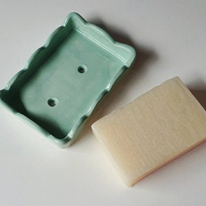 May include: A pale green ceramic soap dish with a wavy edge and two drainage holes. A rectangular bar of light-coloured soap sits beside the dish. The soap and dish are on a white surface.