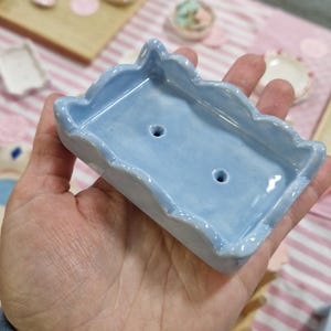 May include: A pale blue ceramic soap dish with a wavy edge and two drainage holes. The rectangular dish is held in a hand, highlighting its glossy finish and handmade quality. Other ceramic items are visible in the background.