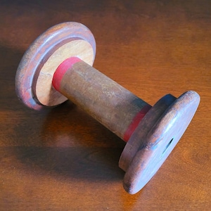 May include: A wooden spool with red and blue painted stripes. The spool is made of wood and has a hole in the center. It is lying on a wooden surface.
