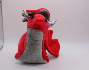 May include: A plush red dragon toy with a gray underbelly, wings, and accents. The dragon has silver horns and small black eyes. The toy is made of soft fabric and is designed to sit upright.