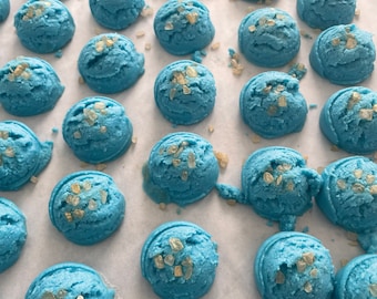 Può includere: Una vista ravvicinata di una superficie bianca con molte bombe da bagno blu. Le bombe da bagno sono rotonde e hanno una consistenza ruvida. Sono ricoperte di piccole scaglie dorate scintillanti.