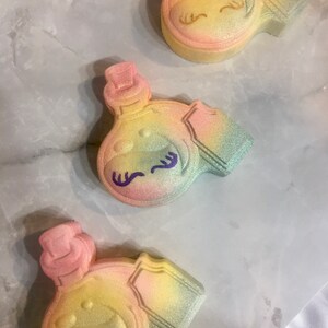 May include: Three pastel-colored bath bombs shaped like bottles with a whimsical face and eyelashes. The bath bombs are decorated with glitter and have a rainbow ombre effect.