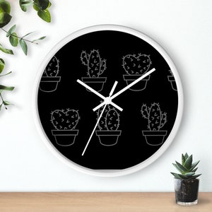 Puede incluir: Reloj blanco y negro con marco blanco y esfera negra. La esfera del reloj tiene un patrón repetitivo de plantas de cactus en macetas. Las manecillas del reloj son blancas.
