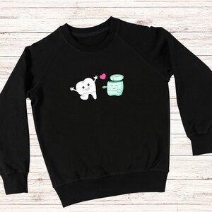 May include: Black sweatshirt with a white tooth and a green floss container graphic. The tooth and floss container are smiling and holding hands. There is a pink heart between them.
