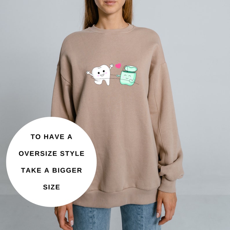 May include: A tan sweatshirt with a cartoon tooth and a container of floss holding hands. The tooth is smiling and the floss container has a happy face. The text "TO HAVE A OVERSIZE STYLE TAKE A BIGGER SIZE" is printed on a white circle in the lower left corner of the image.