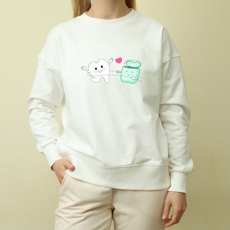 May include: White sweatshirt with a cartoon tooth and a floss container holding hands with a pink heart above them.