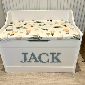 Op de afbeelding: Een witte houten speelgoedkist met een scharnierend deksel en een gewatteerd zitkussen. Het kussen heeft een cr&egrave;mekleurige stof met een patroon van kleine vliegtuigen, heteluchtballonnen en beren. Op de voorkant staat de naam JACK in het blauw.