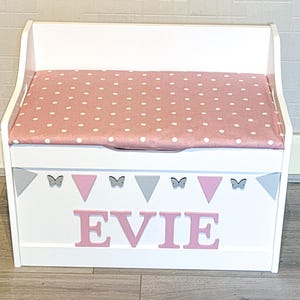 Op de afbeelding: Een witte houten speelgoedkist met een scharnierend deksel voorzien van een roze kussen met witte stippen. Het voorpaneel toont de naam EVIE in roze letters, versierd met een rij afwisselende grijze en roze driehoeken en kleine grijze vlinders.