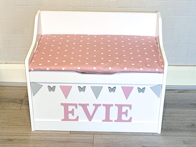Op de afbeelding: Een witte houten speelgoedkist met een scharnierend deksel voorzien van een roze kussen met witte stippen. Het voorpaneel toont de naam EVIE in roze letters, versierd met een rij afwisselende grijze en roze driehoeken en kleine grijze vlinders.