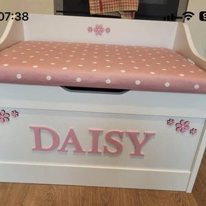 Op de afbeelding: Een witte houten speelgoedkist met een roze gestoffeerde zitting met witte stippen. De voorkant van de kist toont de naam DAISY in grote roze letters, geaccentueerd met kleine roze bloemvormige uitsparingen op de voor- en achterpanelen.
