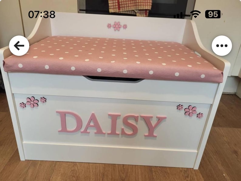 Op de afbeelding: Een witte houten speelgoedkist met een roze gestoffeerde zitting met witte stippen. De voorkant van de kist toont de naam DAISY in grote roze letters, geaccentueerd met kleine roze bloemvormige uitsparingen op de voor- en achterpanelen.
