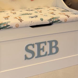 Op de afbeelding: Een witte houten speelgoedkist met de naam SEB in grote grijze letters op de voorzijde. De bovenkant van de kist is een gestoffeerde zitting met een witte bekleding voorzien van illustraties van bruine teddyberen, heteluchtballonnen en vliegtuigen.
