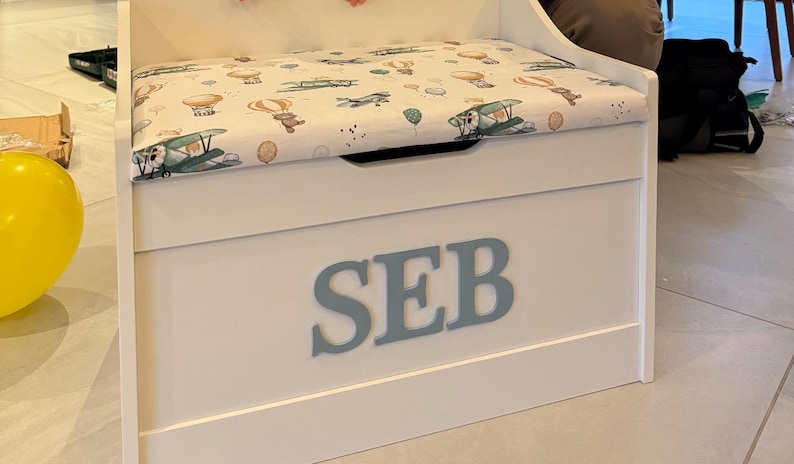 Op de afbeelding: Een witte houten speelgoedkist met de naam SEB in grote grijze letters op de voorzijde. De bovenkant van de kist is een gestoffeerde zitting met een witte bekleding voorzien van illustraties van bruine teddyberen, heteluchtballonnen en vliegtuigen.
