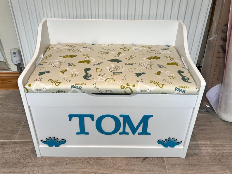 Op de afbeelding: Een witte houten speelgoedkist met een scharnierend deksel voorzien van een beige kussen met dinosauruspatroon. De voorkant van de kist toont de naam TOM in grote blauwe letters, geflankeerd door twee kleine blauwe dinosaurus-silhouetten.
