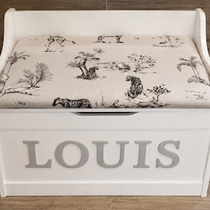 Op de afbeelding: Een witte houten opbergbank met een gestoffeerde zitting in beige stof met een zwart-wit safari-dierenprint. Het voorpaneel van de bank toont de naam LOUIS in grote, verhoogde, lichtgrijze hoofdletters.