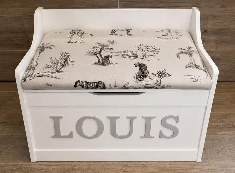 Op de afbeelding: Een witte houten opbergbank met een gestoffeerde zitting in beige stof met een zwart-wit safari-dierenprint. Het voorpaneel van de bank toont de naam LOUIS in grote, verhoogde, lichtgrijze hoofdletters.
