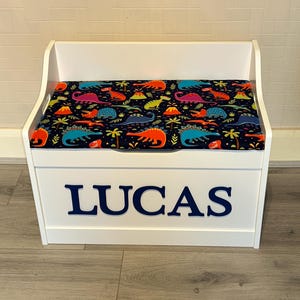 Op de afbeelding: Een witte houten speelgoedkist met een scharnierend deksel voorzien van een kleurrijk dinosauruspatroon op een donkerblauwe achtergrond. De voorzijde van de kist toont de naam LUCAS in grote, dikke, donkerblauwe hoofdletters in het midden.