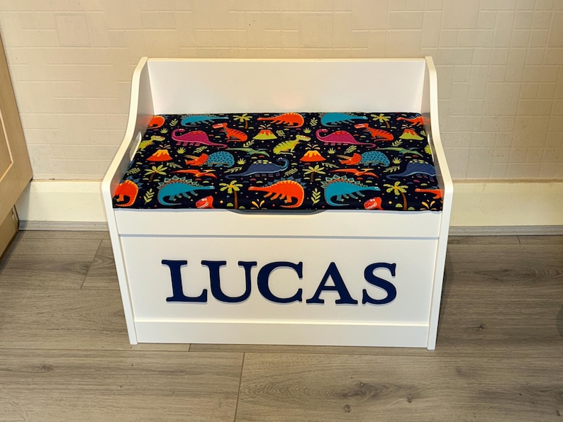 Op de afbeelding: Een witte houten speelgoedkist met een scharnierend deksel voorzien van een kleurrijk dinosauruspatroon op een donkerblauwe achtergrond. De voorzijde van de kist toont de naam LUCAS in grote, dikke, donkerblauwe hoofdletters in het midden.