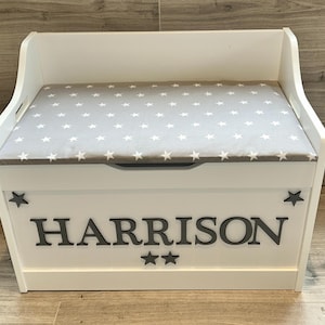 Op de afbeelding: Een witte houten speelgoedkist met een grijs gestoffeerd zitkussen met een wit sterrenpatroon. Het voorpaneel toont de naam HARRISON in grote grijze hoofdletters, geflankeerd door twee kleine grijze sterren aan de zijkanten en twee onder de naam.