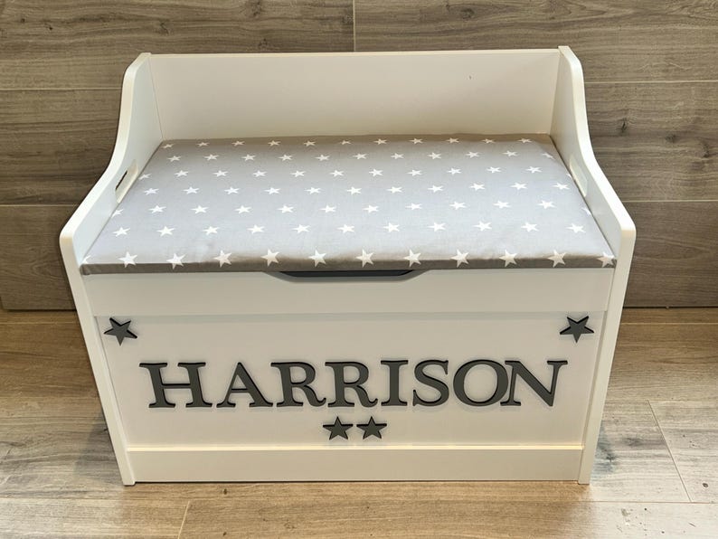 Op de afbeelding: Een witte houten speelgoedkist met een grijs gestoffeerd zitkussen met een wit sterrenpatroon. Het voorpaneel toont de naam HARRISON in grote grijze hoofdletters, geflankeerd door twee kleine grijze sterren aan de zijkanten en twee onder de naam.