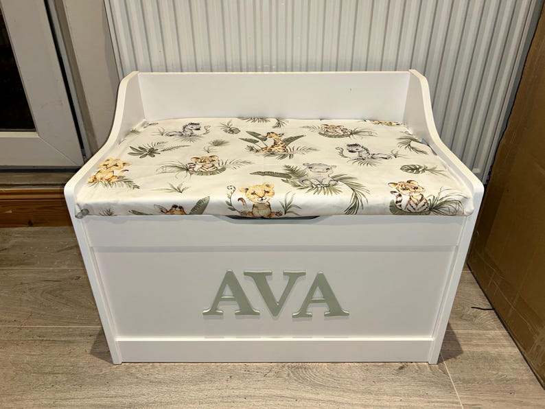 Op de afbeelding: Een witte houten speelgoedkist met een gewatteerd zitkussen met een jungle-dierenprint. Op de voorkant van de kist staat de naam AVA in grote, lichte hoofdletters. Het ontwerp is voorzien van een uitgesneden handgreep om de kist te openen.
