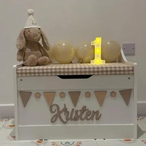 Op de afbeelding: Een witte houten speelgoedkist met een beige en wit geblokt kussen bovenop. De voorkant heeft een houten uitgesneden naam "Kristen" onder een rij driehoekige vlaggetjes en bloemvormen. Een pluche konijn met een feesthoedje zit bovenop de kist.