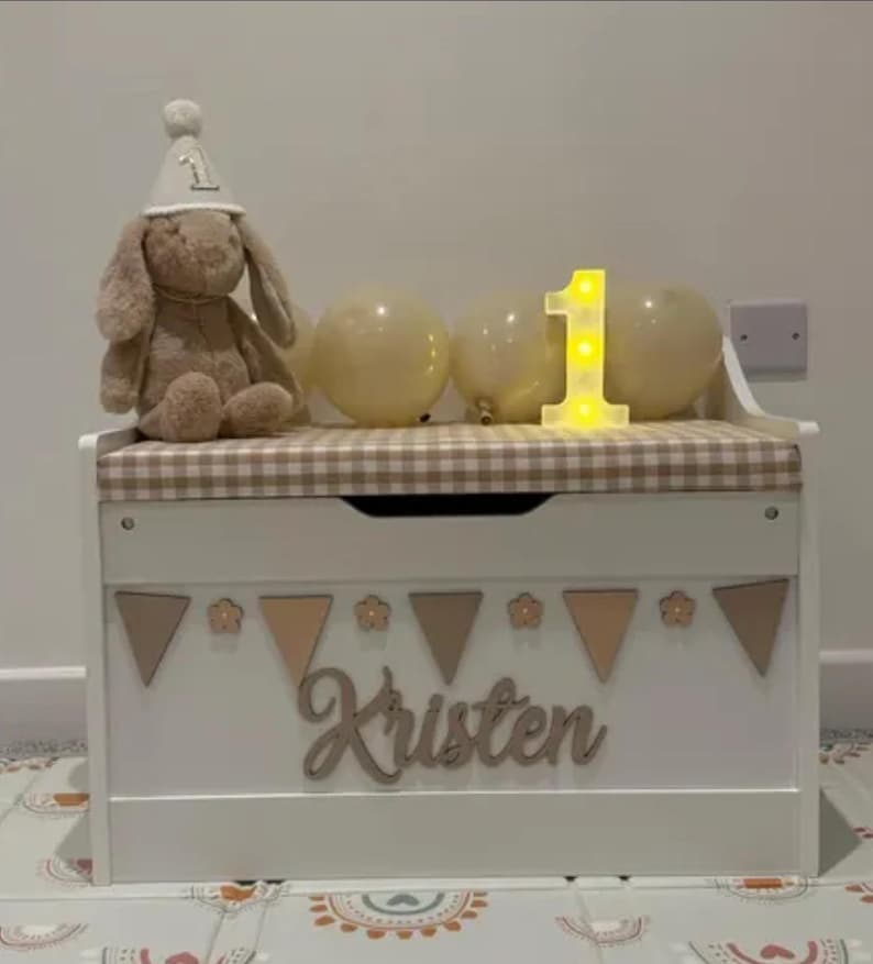 Op de afbeelding: Een witte houten speelgoedkist met een beige en wit geblokt kussen bovenop. De voorkant heeft een houten uitgesneden naam "Kristen" onder een rij driehoekige vlaggetjes en bloemvormen. Een pluche konijn met een feesthoedje zit bovenop de kist.