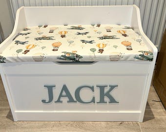 Op de afbeelding: Een witte houten speelgoedkist met een gewatteerd deksel met een patroon van vliegtuigen en luchtballonnen. De naam "JACK" staat in grijze letters op de voorkant. De kist is ontworpen voor opslag.