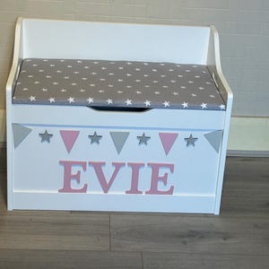 Op de afbeelding: Een witte houten speelgoedkist met een grijs kussen voorzien van een wit sterrenpatroon. Het voorpaneel is versierd met een slinger van grijze sterren en roze driehoeken, en de naam EVIE staat in grote, roze blokletters op de voorzijde.