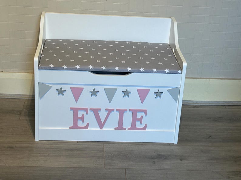 Op de afbeelding: Een witte houten speelgoedkist met een grijs kussen voorzien van een wit sterrenpatroon. Het voorpaneel is versierd met een slinger van grijze sterren en roze driehoeken, en de naam EVIE staat in grote, roze blokletters op de voorzijde.