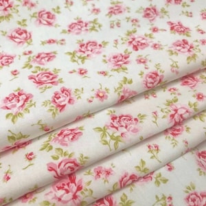 Op de afbeelding: Een gevouwen lengte witte stof met een herhalend patroon van kleine roze rozen en lichtgroene bladeren. Het textiel vertoont een zachte textuur met meerdere lagen zichtbaar in de plooien, wat de bloemenprint over het oppervlak laat zien.