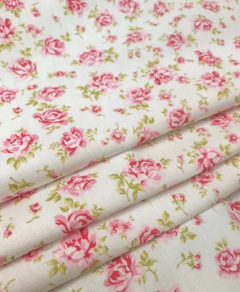 Op de afbeelding: Een gevouwen lengte witte stof met een herhalend patroon van kleine roze rozen en lichtgroene bladeren. Het textiel vertoont een zachte textuur met meerdere lagen zichtbaar in de plooien, wat de bloemenprint over het oppervlak laat zien.