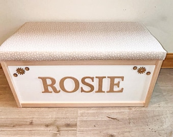 Op de afbeelding: Een rechthoekige houten opbergkist met een wit voorpaneel waarop de naam ROSIE in grote gouden letters staat. Het deksel is bekleed met een lichtbeige stof met een klein wit bloemenpatroon. Kleine gouden bloemaccenten zijn geplaatst in de bovenhoeken van het voorpaneel.