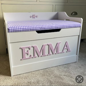 Op de afbeelding: Een witte houten speelgoedkist met een paars-wit geblokt stoffen kussen aan de bovenkant. De voorkant van de kist toont de naam EMMA in grote, lichtroze, driedimensionale letters. Een kleine paarse bloemdecoratie is gecentreerd op het achterpaneel.