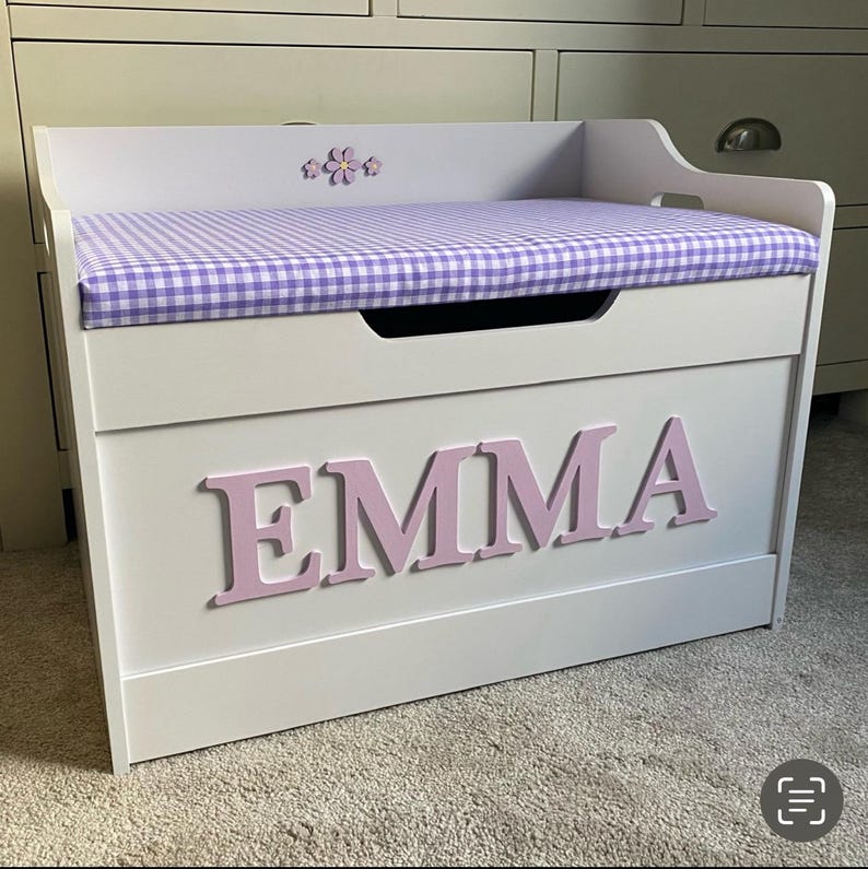 Op de afbeelding: Een witte houten speelgoedkist met een paars-wit geblokt stoffen kussen aan de bovenkant. De voorkant van de kist toont de naam EMMA in grote, lichtroze, driedimensionale letters. Een kleine paarse bloemdecoratie is gecentreerd op het achterpaneel.