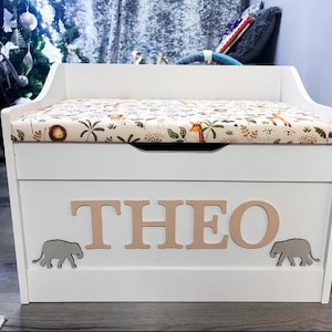 Op de afbeelding: Een witte houten speelgoedkist met een gestoffeerde zitting en een beige stoffen hoes met een print van oerwouddieren. Op de voorkant van de kist staat de naam THEO in grote lichte letters, geflankeerd door twee kleine grijze olifantjes.