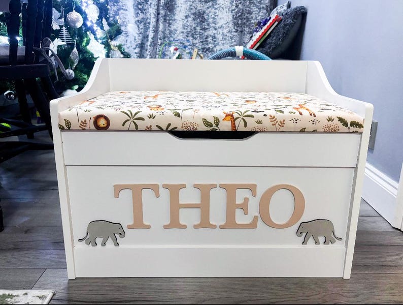 Op de afbeelding: Een witte houten speelgoedkist met een gestoffeerde zitting en een beige stoffen hoes met een print van oerwouddieren. Op de voorkant van de kist staat de naam THEO in grote lichte letters, geflankeerd door twee kleine grijze olifantjes.