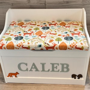 Op de afbeelding: Een witte houten speelgoedkist met een gestoffeerde zitting voorzien van een kleurrijk bospatroon met oranje vossen, konijnen en bomen. De voorkant van de kist toont de naam CALEB in grote grijze letters, met een kleine oranje vos en bruine paddenstoelen.