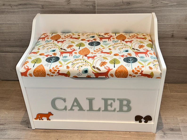 Op de afbeelding: Een witte houten speelgoedkist met een gestoffeerde zitting voorzien van een kleurrijk bospatroon met oranje vossen, konijnen en bomen. De voorkant van de kist toont de naam CALEB in grote grijze letters, met een kleine oranje vos en bruine paddenstoelen.
