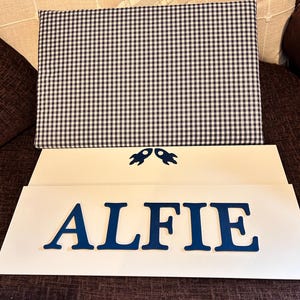 Op de afbeelding: Een rechthoekig stoffen paneel met een marineblauw en wit gingham-patroon boven twee witte rechthoekige borden. Het bovenste bord bevat twee kleine marineblauwe raketpictogrammen en het onderste bord toont de naam ALFIE in marineblauw.