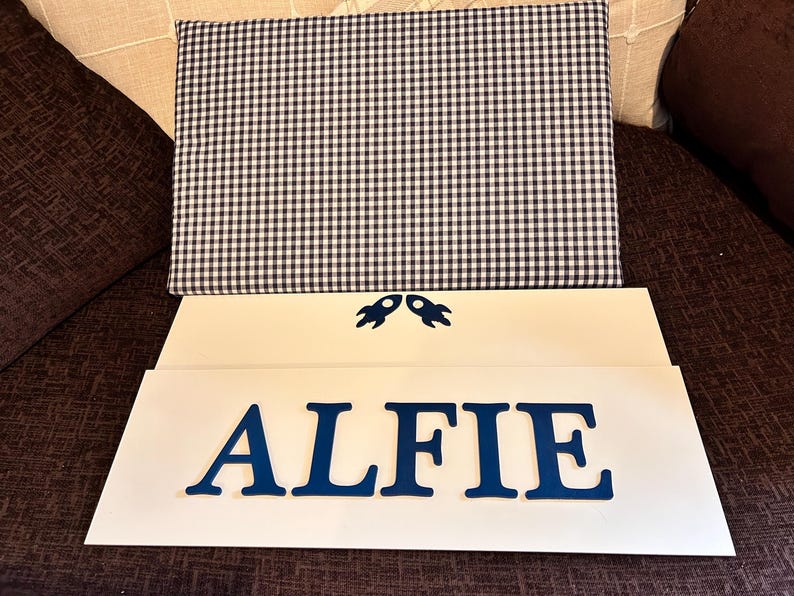 Op de afbeelding: Een rechthoekig stoffen paneel met een marineblauw en wit gingham-patroon boven twee witte rechthoekige borden. Het bovenste bord bevat twee kleine marineblauwe raketpictogrammen en het onderste bord toont de naam ALFIE in marineblauw.