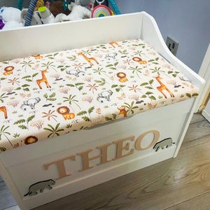 Op de afbeelding: Een witte houten speelgoedkist met een gestoffeerd zitkussen voorzien van een jungle-dierenprint met giraffen, leeuwen en olifanten. De voorzijde van de kist toont de naam THEO in verhoogde houten letters, geflankeerd door twee kleine olifantjes.