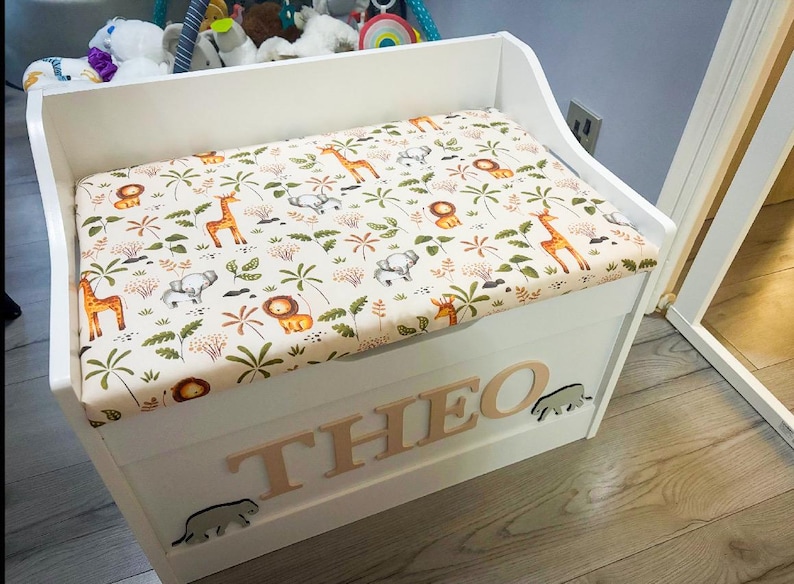 Op de afbeelding: Een witte houten speelgoedkist met een gestoffeerd zitkussen voorzien van een jungle-dierenprint met giraffen, leeuwen en olifanten. De voorzijde van de kist toont de naam THEO in verhoogde houten letters, geflankeerd door twee kleine olifantjes.