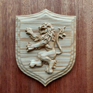 May include: A wooden shield with a carved lion in relief. The lion is facing right and has a long tail. The shield is a light brown colour and has a smooth surface.