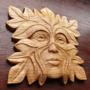 May include: A wooden carving of a Green Man's face, with leaves and branches forming the face and hair. The carving is detailed and realistic, with a warm brown color.