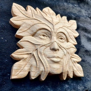 May include: A wooden carving of a Green Man face, with leaves and branches forming the face and hair. The carving is unfinished, with the wood still showing its natural grain.