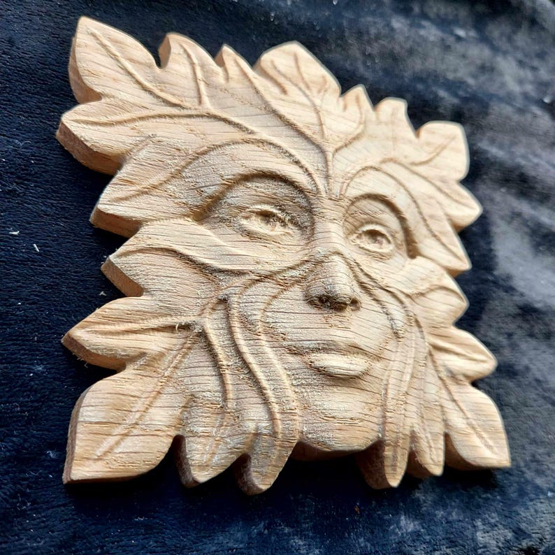 May include: A wooden carving of a Green Man face, with leaves and branches forming the face and hair. The carving is unfinished, with the wood still showing its natural grain.