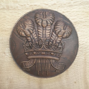 May include: A dark brown wooden plaque with a carved image of three feathers and a crown. The plaque has the text "ICH DIEN" on a banner below the crown.